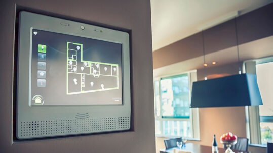 Wandmontiertes Smart-Home-Display zeigt den Grundriss einer Wohnung mit vernetzten Geräten und Lichtsteuerung in einem modernen Wohnraum. | Immobilienbewertung